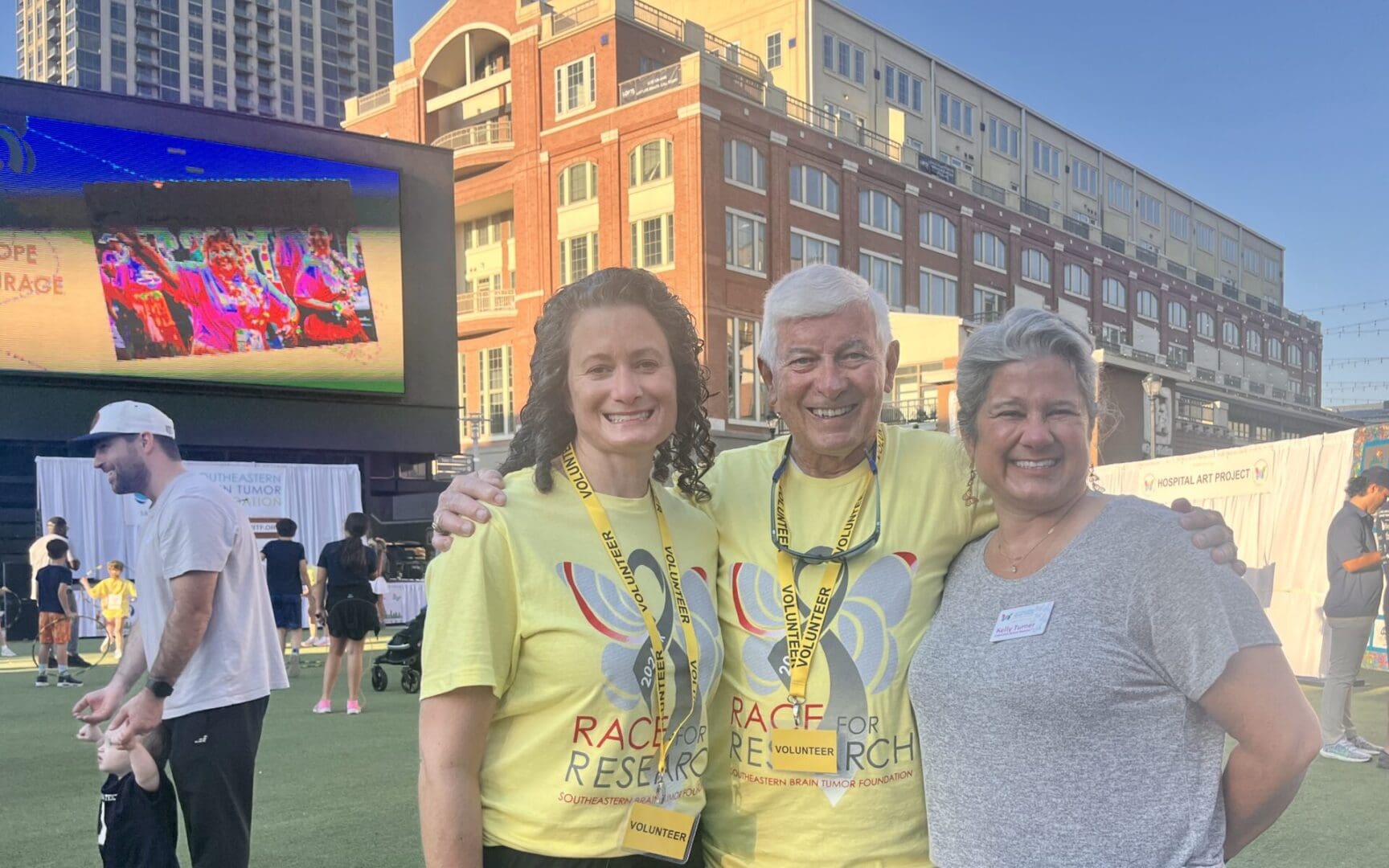 Three smiling women wearing matching yellow event t-shirts outdoors.