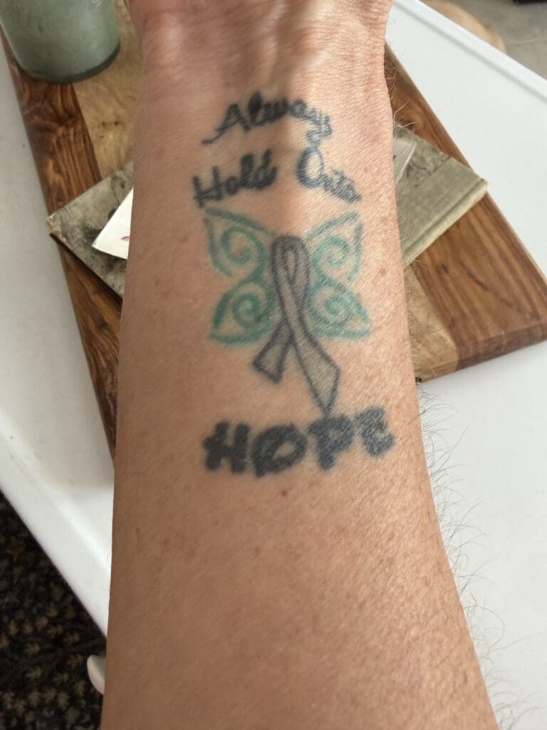 Forearm tattoo reading Always Hold On To Hope around a teal butterfly and a gray awareness ribbon